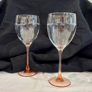 Puede incluir: Dos copas de vino transparentes con un delicado diseño de hojas grabado cerca del borde. Los tallos y las bases son de color melocotón claro. Las copas están sobre un fondo oscuro.