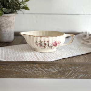 Könnte beinhalten: Cremefarbene Sauciere mit einem Blumendesign in Rosa und Grün. Der Rand und der Griff sind goldfarben verziert. Das Boot hat einen Ausguss und eine gerippte Außenseite.