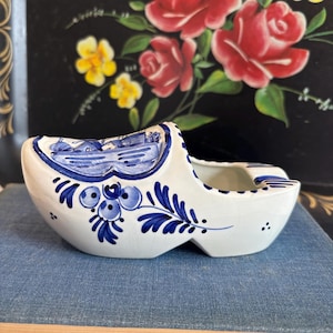 May include: White ceramic clog-shaped ashtray with blue floral and landscape designs. The ashtray features a classic Dutch design with a detailed village scene and decorative accents. A unique and functional decorative piece.
