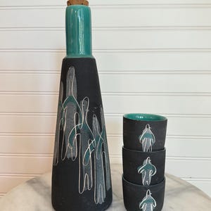 Puede incluir: Una jarra de cerámica negra con tapa y tapón de corcho turquesa, con figuras abstractas blancas y turquesas. Junto a ella, tres tazas negras con interiores turquesas y figuras a juego.