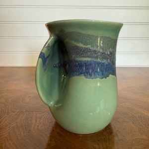 Puede incluir: Una taza de cerámica hecha a mano con un asa única. La taza presenta un degradado de colores, que va desde una base verde claro hasta un borde verde oscuro y azul. El diseño de la taza es orgánico y el asa está integrada en el cuerpo.