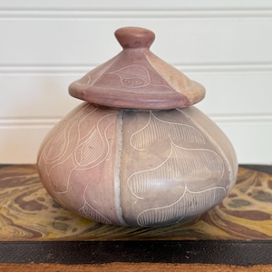 May include: A stone jar with a lid, featuring etched leaf designs. The jar has a rounded body and a conical lid. The colour scheme includes pink, tan, and grey tones. The jar is likely used for storage or display, and is measured in centimetres.