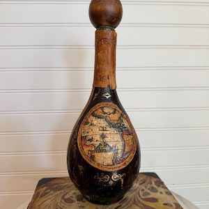 Könnte beinhalten: Eine dunkel gefärbte Vintage-Flasche mit einem Holzstopfen. Die Flasche zeigt ein detailliertes Kartendesign auf der Vorderseite und steht auf einem Buch. Der Flaschenhals ist mit gealtertem Leder umwickelt.