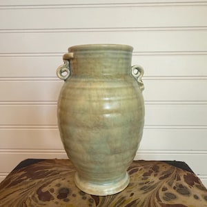 Może przedstawiać: Wysoki, jasnozielony ceramiczny wazon z dwoma uchwytami w kształcie pętli. Wazon ma szeroki korpus i lekko rozszerzoną podstawę. Powierzchnia ma marmurkowy wygląd z odcieniami. Przedmiot dekoracyjny.