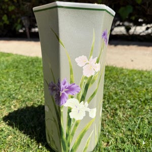 May include: A tall, hexagonal vase in a muted green colour, decorated with painted iris flowers in shades of purple, white, and pink. The vase has green trim and long green leaves. The vase is standing on a grassy surface.