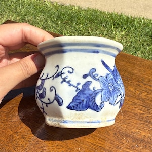 Könnte beinhalten: Ein kleiner, weißer Keramiktopf mit blauen Blumen- und Blattmotiven. Der Topf hat einen leicht ausgestellten Rand und einen abgerundeten Körper. Die blauen Designs sind handgemalt. Der Topf wird von einer Hand gehalten.