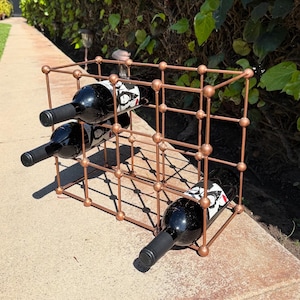 May include: A copper-colored metal wine rack holding four bottles of red wine. The rack has a grid-like design with spherical joints. The wine bottles are black with white and red labels.
