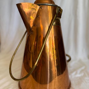 May include: A copper tea kettle with a conical body and a spout. It features a brass handle and a lid with a small knob. The copper surface reflects light, creating a warm glow. The kettle is likely vintage, a classic kitchenware item.