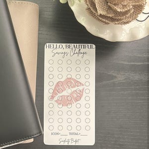 以下が含まれることがあります： 「Hello, Beautiful Savings Challenge」の文字とピンクの唇のプリントが入った白い貯蓄チャレンジカード。貯蓄を追跡するための複数の円があります。カードは暗い表面にあります。
