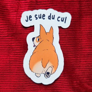 Może przedstawiać: Naklejka przedstawiająca kreskówkowego corgi z tyłu, z pomarańczowym futrem i czarnymi poduszkami łap. Naklejka ma tekst "je sue du cul" w kolorze niebieskim na białym tle. Naklejka znajduje się na czerwonej, teksturowanej powierzchni.