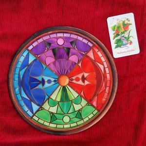 Pode incluir: Um prato decorativo redondo e colorido com um design de vitral. O prato apresenta segmentos azuis, verdes, vermelhos, roxos e laranja. Um pequeno cartão com uma ilustração de morangos está ao lado do prato. Borda marrom.