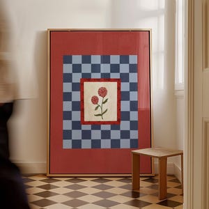 May include: A framed artwork featuring a floral design. The artwork has a red border, a blue and gray checkered pattern, and a central square with two red flowers. The frame is gold, and the artwork is displayed in a room with a checkered floor.
