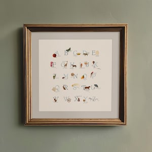 May include: Framed artwork featuring an illustrated alphabet with corresponding images. The gold frame surrounds a cream-colored print with colorful drawings of an apple, butterfly, crown, dog, and egg, among other objects.