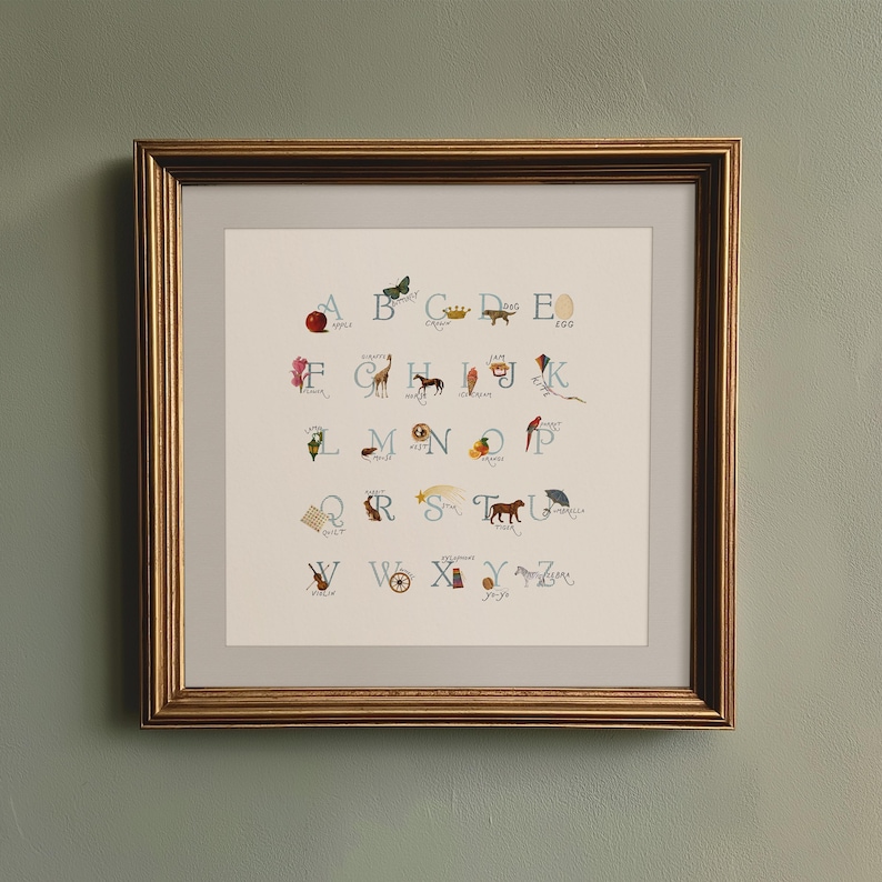 May include: Framed artwork featuring an illustrated alphabet with corresponding images. The gold frame surrounds a cream-colored print with colorful drawings of an apple, butterfly, crown, dog, and egg, among other objects.