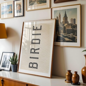 Puede incluir: Una habitaci&oacute;n con arte enmarcado y decoraci&oacute;n. Una gran impresi&oacute;n enmarcada con la palabra "BIRDIE" en letras negras en bloque se apoya contra la pared. Tambi&eacute;n se ven otras im&aacute;genes enmarcadas y una l&aacute;mpara. Un mueble de madera y jarrones decorativos completan la escena.