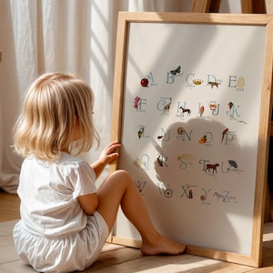 May include: A framed alphabet chart with colorful illustrations and corresponding letters. The artwork is in a light wooden frame and is leaning against a wooden easel. A young child is sitting on the floor, looking at the chart.