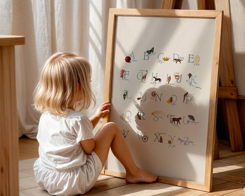 May include: A framed alphabet chart with colorful illustrations and corresponding letters. The artwork is in a light wooden frame and is leaning against a wooden easel. A young child is sitting on the floor, looking at the chart.