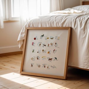 May include: A framed alphabet art print with a wooden frame. Each letter is illustrated with a corresponding image, such as a butterfly for B and a giraffe for G. The artwork is displayed on a wooden floor, leaning against a bed.