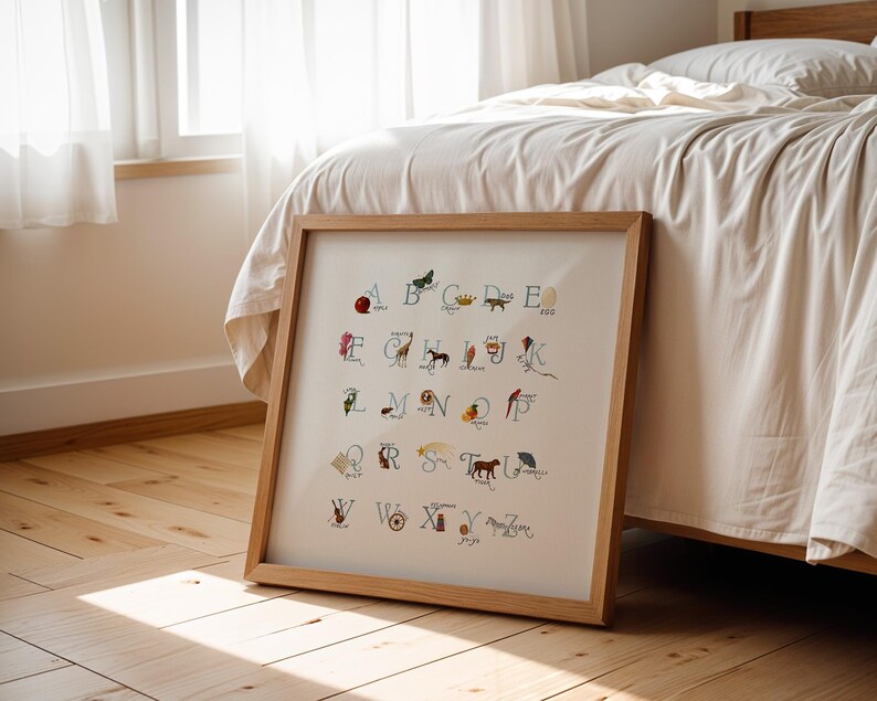 May include: A framed alphabet art print with a wooden frame. Each letter is illustrated with a corresponding image, such as a butterfly for B and a giraffe for G. The artwork is displayed on a wooden floor, leaning against a bed.