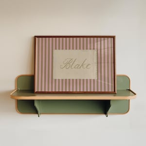 May include: A rectangular picture frame with a dark wood finish containing a print with vertical pink and tan stripes and a central beige rectangle featuring the name Blake in cursive script, resting on a green wall-mounted shelf with light trim.
