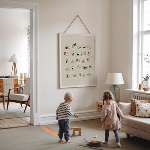 May include: A framed alphabet chart with colorful illustrations of animals and objects, hanging on a white wall. The artwork is displayed in a bright, airy room with children playing on the floor.
