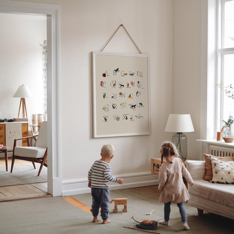 May include: A framed alphabet chart with colorful illustrations of animals and objects, hanging on a white wall. The artwork is displayed in a bright, airy room with children playing on the floor.