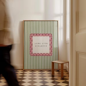May include: A framed art print with a gold frame, featuring a green and white striped background. The print has a central square with the words "LONG LIVE GIRLHOOD" in black text, framed by a pink floral border. A small wooden stool sits nearby.
