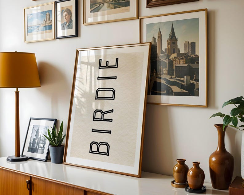 May include: A room with framed art and decor. A large framed print with the word "BIRDIE" in black block letters leans against the wall. Other framed pictures and a lamp are also visible. A wooden cabinet and decorative vases complete the scene.