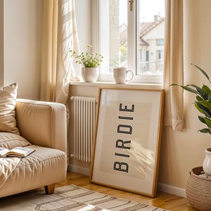 Puede incluir: Una impresi&oacute;n enmarcada con la palabra "BIRDIE" en letras negras se apoya contra una pared en una habitaci&oacute;n bien iluminada. Tambi&eacute;n se ven un sof&aacute; beige, una planta en maceta y una taza en un alf&eacute;izar. La habitaci&oacute;n tiene una paleta de colores neutros.