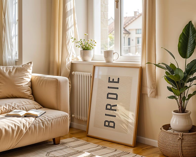 Puede incluir: Una impresi&oacute;n enmarcada con la palabra "BIRDIE" en letras negras se apoya contra una pared en una habitaci&oacute;n bien iluminada. Tambi&eacute;n se ven un sof&aacute; beige, una planta en maceta y una taza en un alf&eacute;izar. La habitaci&oacute;n tiene una paleta de colores neutros.