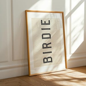 Puede incluir: Una impresi&oacute;n art&iacute;stica enmarcada con la palabra "BIRDIE" en letras negras delineadas. La impresi&oacute;n tiene un fondo beige y se muestra en un marco de madera marr&oacute;n claro. La obra de arte est&aacute; apoyada contra una pared blanca y un suelo de madera.