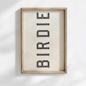 Puede incluir: Impresi&oacute;n enmarcada con la palabra "BIRDIE" en letras negras en negrita y delineadas. El texto est&aacute; orientado verticalmente sobre un fondo color crema. El marco es de color dorado.