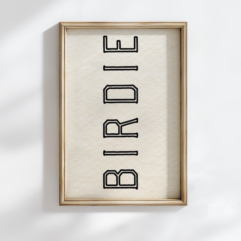Puede incluir: Impresi&oacute;n enmarcada con la palabra "BIRDIE" en letras negras en negrita y delineadas. El texto est&aacute; orientado verticalmente sobre un fondo color crema. El marco es de color dorado.
