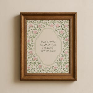May include: A framed print with a floral pattern and the words "THIS LITTLE LIGHT OF MINE, I'M GONNA LET IT SHINE." The frame is dark brown. The background has pink roses and green leaves.