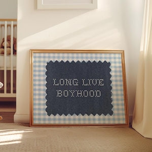 Può includere: Un'opera d'arte incorniciata con la scritta "LONG LIVE BOYHOOD" ricamata su un tessuto blu navy. La cornice ha una finitura dorata e uno sfondo a quadretti azzurri. Un cavallo a dondolo bianco e una culla sono sullo sfondo.