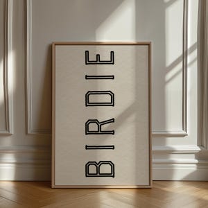 Puede incluir: Una impresi&oacute;n enmarcada con la palabra "BIRDIE" en letras negras en negrita y delineadas. El texto est&aacute; centrado en un fondo beige, dentro de un marco de madera clara. La obra de arte se exhibe contra una pared blanca.