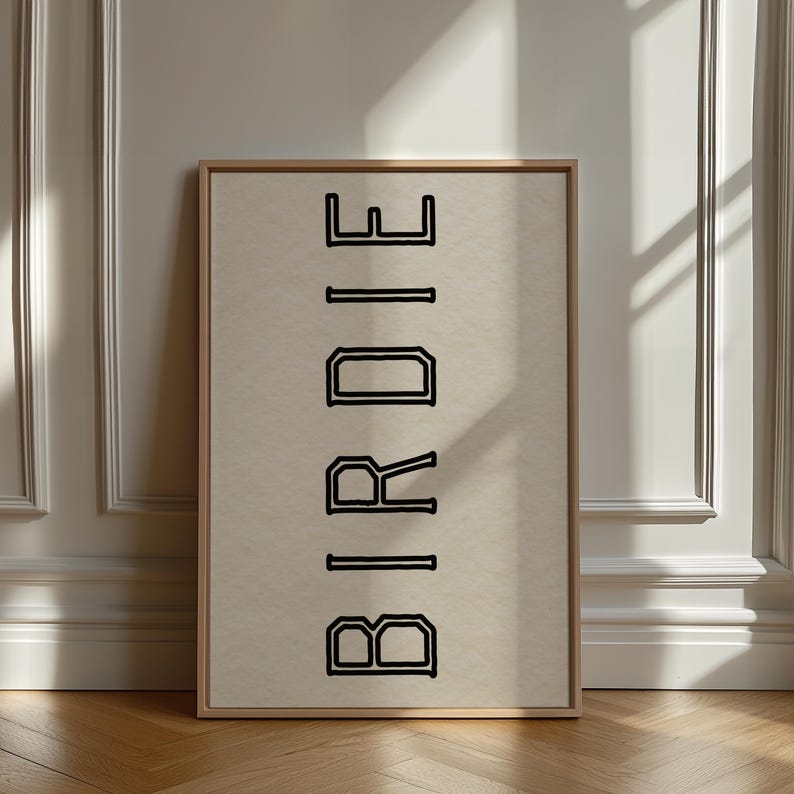 Puede incluir: Una impresi&oacute;n enmarcada con la palabra "BIRDIE" en letras negras en negrita y delineadas. El texto est&aacute; centrado en un fondo beige, dentro de un marco de madera clara. La obra de arte se exhibe contra una pared blanca.