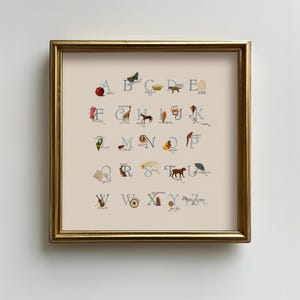 May include: A framed alphabet art print featuring whimsical illustrations for each letter. The artwork includes a red apple, a butterfly, a crown, a dog, and an egg. The print is in a gold frame and is displayed on a white wall.