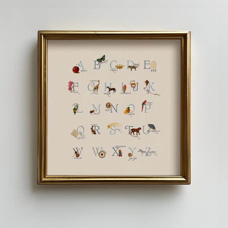 May include: A framed alphabet art print featuring whimsical illustrations for each letter. The artwork includes a red apple, a butterfly, a crown, a dog, and an egg. The print is in a gold frame and is displayed on a white wall.