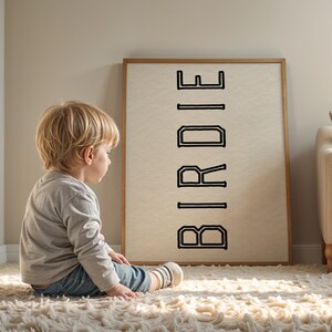 Puede incluir: Una obra de arte enmarcada con la palabra "BIRDIE" en letras negras. Un ni&ntilde;o peque&ntilde;o est&aacute; sentado en una alfombra blanca y esponjosa, mirando la obra de arte. El marco es de color marr&oacute;n claro y el fondo es beige neutro.