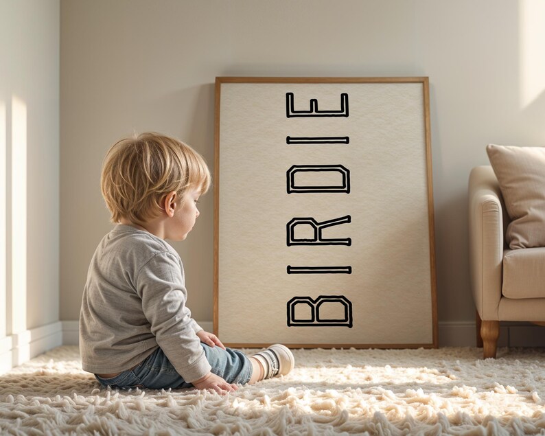 Puede incluir: Una obra de arte enmarcada con la palabra "BIRDIE" en letras negras. Un ni&ntilde;o peque&ntilde;o est&aacute; sentado en una alfombra blanca y esponjosa, mirando la obra de arte. El marco es de color marr&oacute;n claro y el fondo es beige neutro.