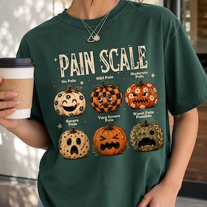 May include: A dark green t-shirt featuring a graphic titled PAIN SCALE with six pumpkins displaying different facial expressions. The pumpkins are labeled No Pain, Mild Pain, Moderate Pain, Severe Pain, Very Severe Pain, and Worst Pain Possible.