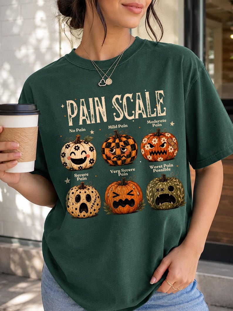 May include: A dark green t-shirt featuring a graphic titled PAIN SCALE with six pumpkins displaying different facial expressions. The pumpkins are labeled No Pain, Mild Pain, Moderate Pain, Severe Pain, Very Severe Pain, and Worst Pain Possible.
