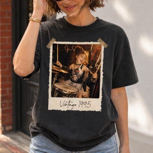 以下が含まれることがあります： ヴィンテージフォトプリントが施された黒いTシャツ。写真にはドラムを演奏する若い女の子が写っており、その下に「Vintage 1995」の文字が書かれています。Tシャツは、クラシックなクルーネックと半袖です。