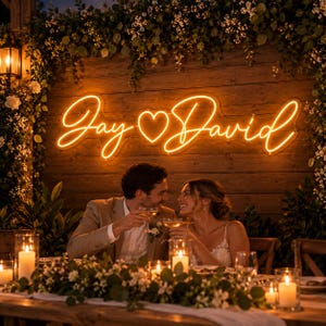 May include: A custom neon sign featuring the names Jay and David in a glowing orange cursive script with a heart symbol in the center, mounted on a rustic wooden wall backdrop decorated with lush green foliage and small white flowers.