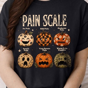 May include: A black t-shirt featuring a graphic titled PAIN SCALE with six pumpkins showing different facial expressions. The pumpkins represent levels from No Pain to Worst Pain Possible, decorated with flowers, checkers, and bandages.