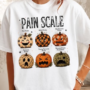 May include: A white t-shirt featuring a graphic titled PAIN SCALE with six illustrated pumpkins showing different facial expressions. The pumpkins are labeled No Pain, Mild Pain, Moderate Pain, Severe Pain, Very Severe Pain, and Worst Pain Possible.