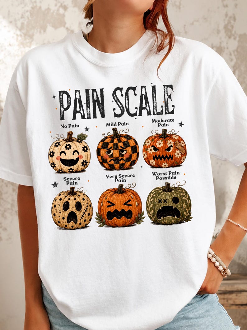 May include: A white t-shirt featuring a graphic titled PAIN SCALE with six illustrated pumpkins showing different facial expressions. The pumpkins are labeled No Pain, Mild Pain, Moderate Pain, Severe Pain, Very Severe Pain, and Worst Pain Possible.