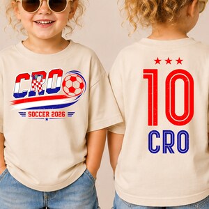 Może przedstawiać: Beżowy t-shirt z grafiką z przodu zawierającą napis CRO, piłkę nożną i SOCCER 2026. Z tyłu koszulki znajduje się czerwona liczba 10, trzy czerwone gwiazdki nad nią oraz niebieski napis CRO umieszczony pod liczbą w pogrubionej czcionce.