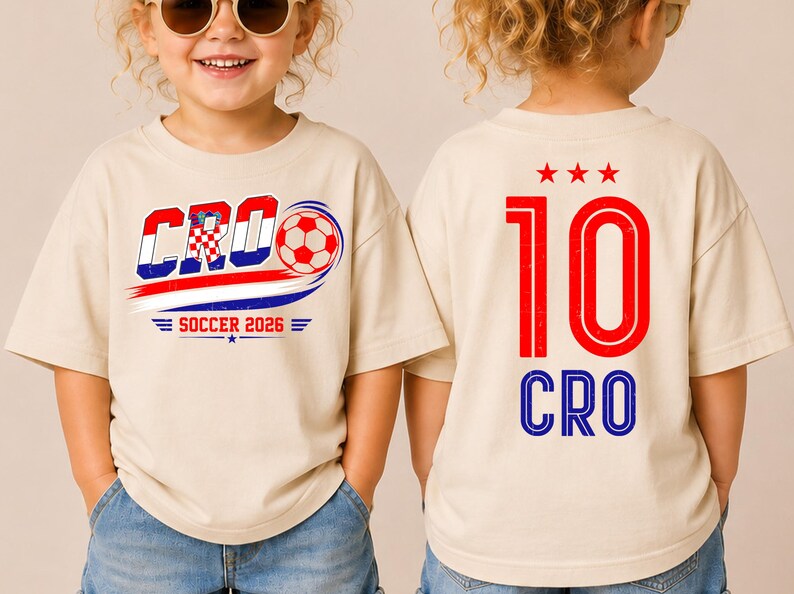 Może przedstawiać: Beżowy t-shirt z grafiką z przodu zawierającą napis CRO, piłkę nożną i SOCCER 2026. Z tyłu koszulki znajduje się czerwona liczba 10, trzy czerwone gwiazdki nad nią oraz niebieski napis CRO umieszczony pod liczbą w pogrubionej czcionce.
